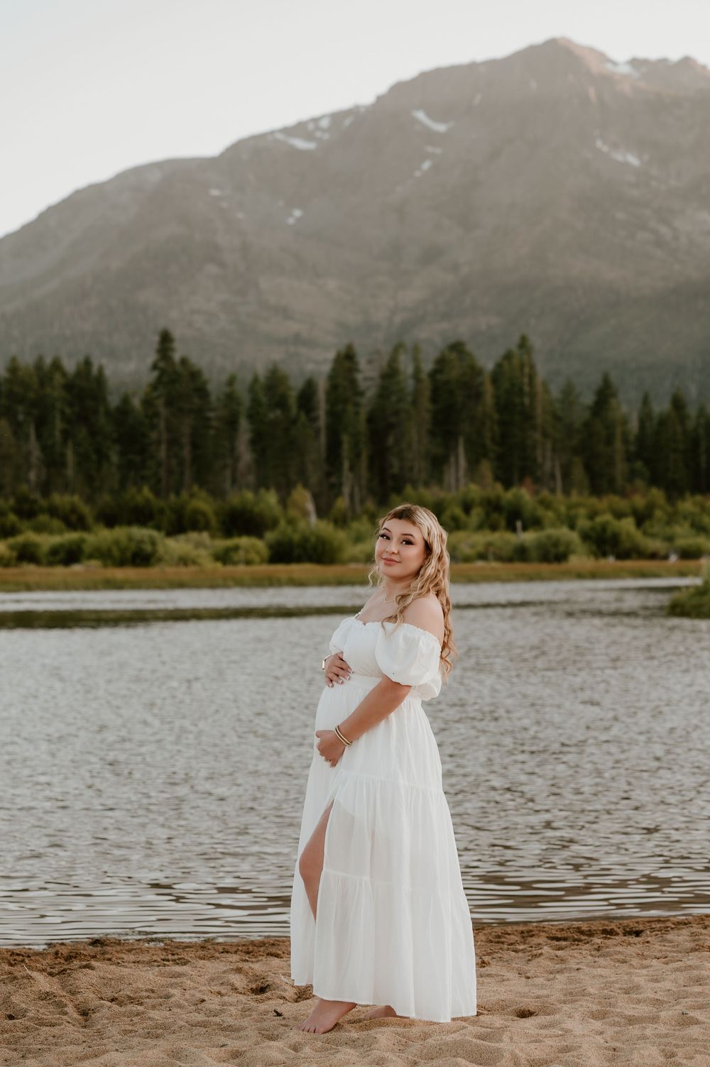 Pregnant woman in white off-shoulder maxi dress standing on beach with mountain lake scenery behind her.