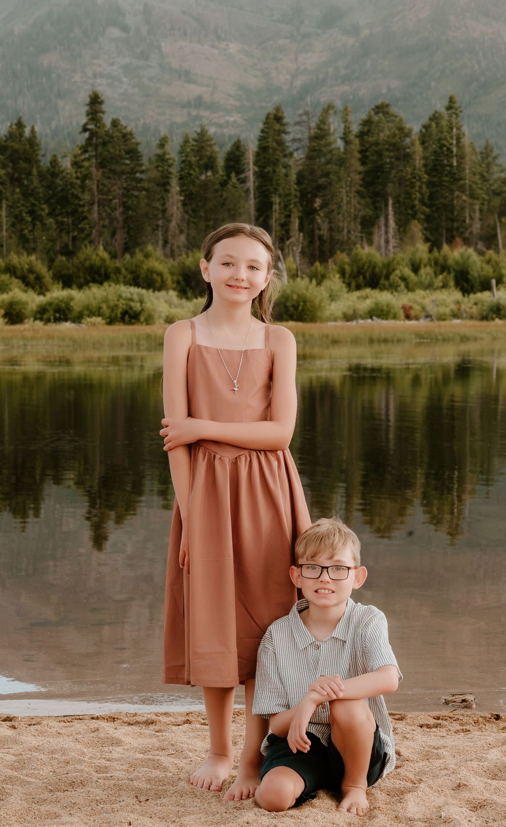 Two children pose by a mountain lake, girl in rust dress standing, boy in polo shirt sitting on sandy shore.