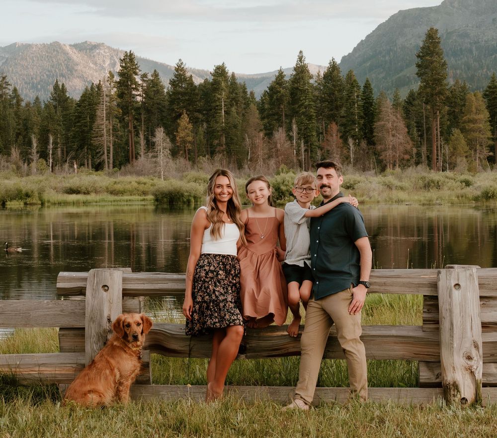 Family of three with a dog posing on a wooden fence near a mountain lake surrounded by tall pine trees.