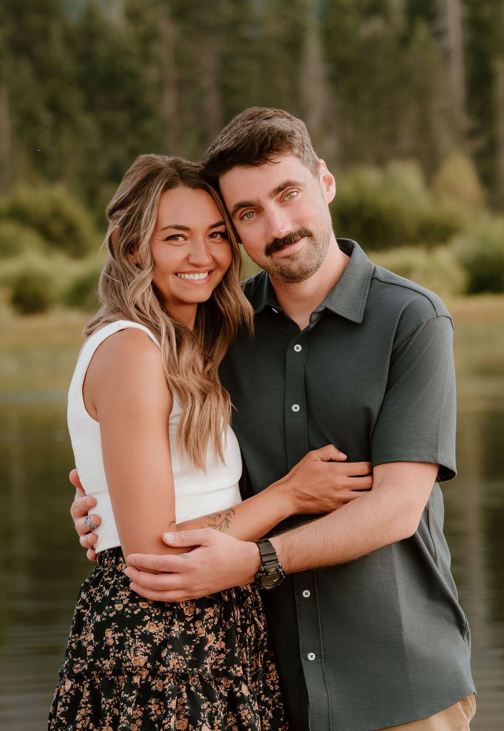 A smiling couple embraces outdoors near a lake surrounded by pine trees, wearing casual summer attire.
