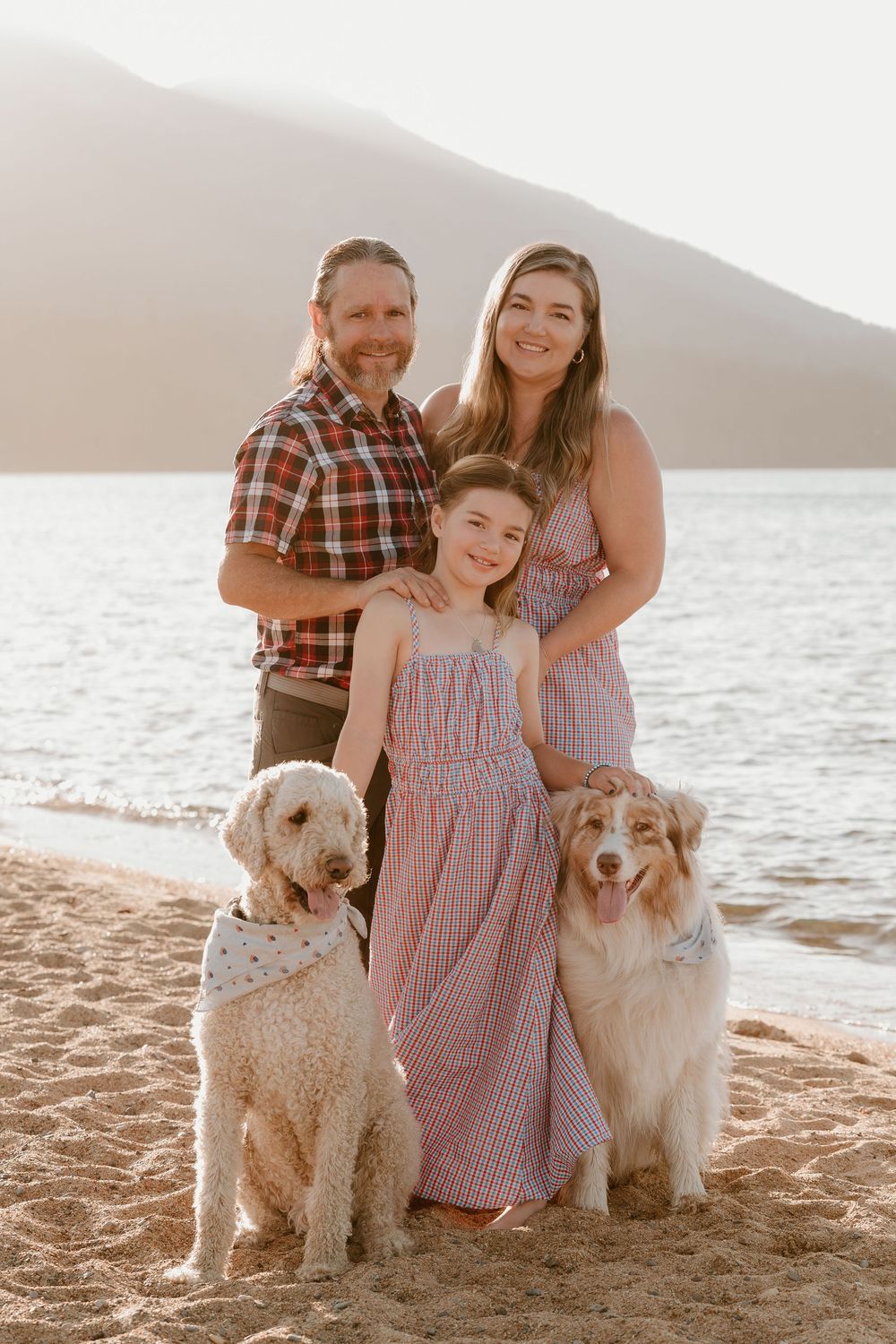 Family of three with two dogs posing on a sandy beach at sunset with mountains in the background.