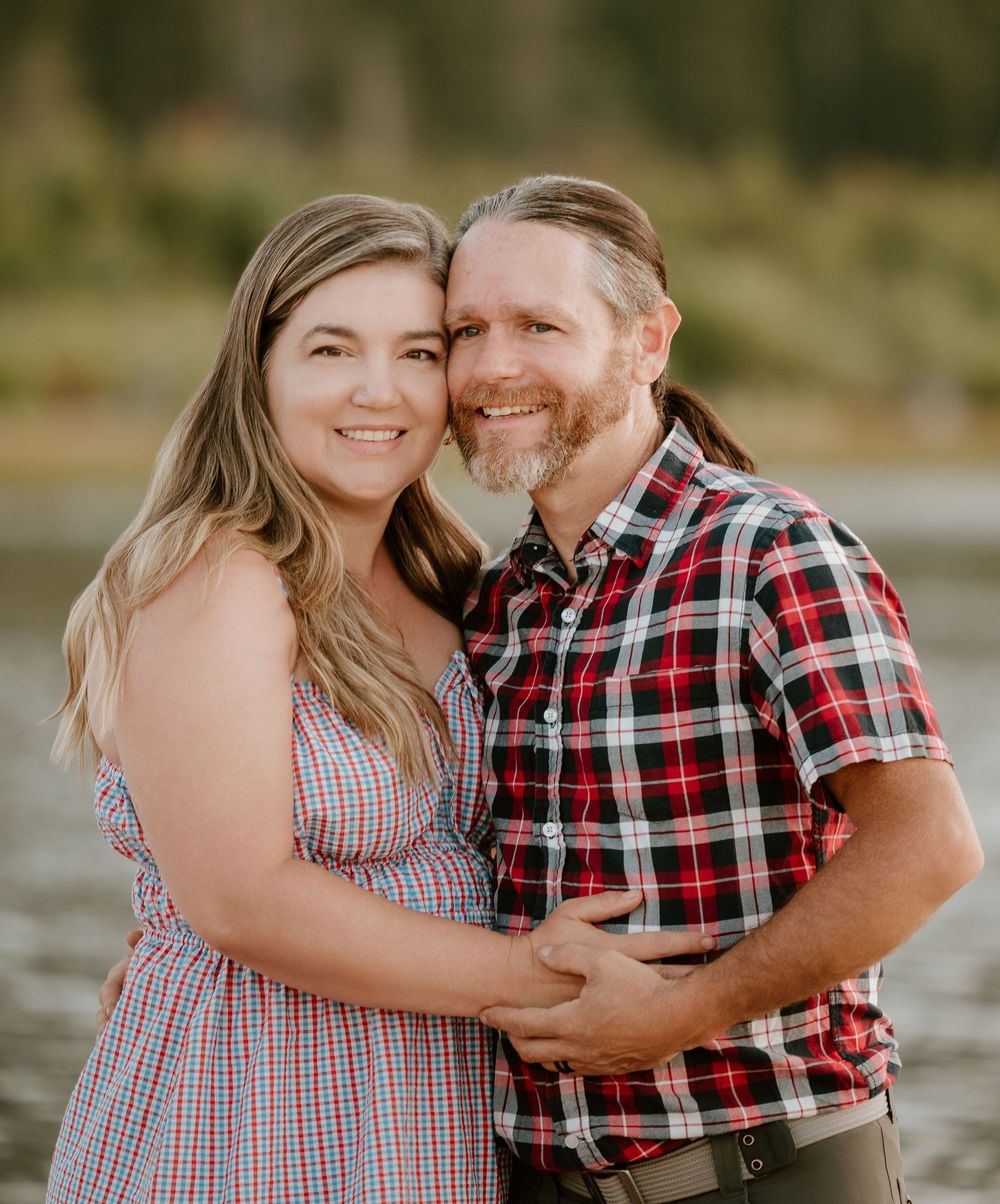 A smiling couple embraces outdoors near a lake, woman in floral dress, man in red plaid shirt with long hair and beard.
