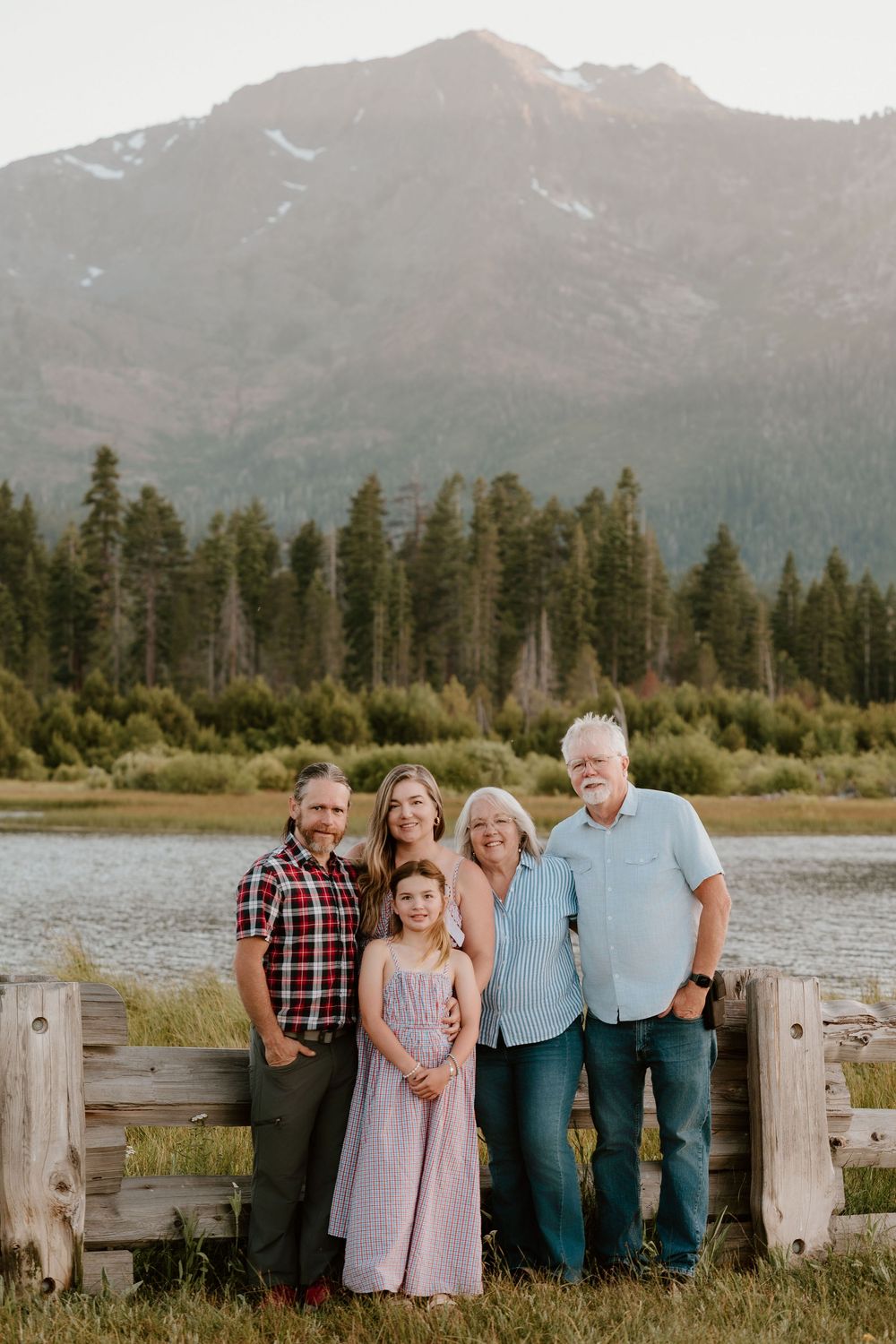 Family of five posing near a mountain lake with pine trees and snow-capped peaks in the background.