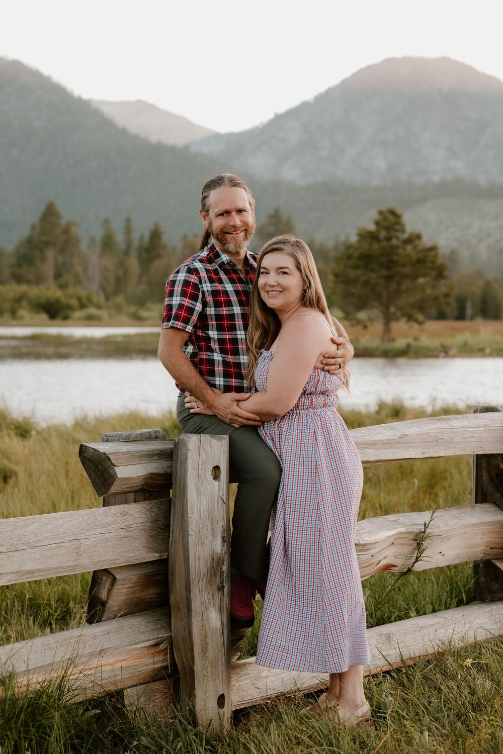 Couple embracing by a wooden fence with mountain lake backdrop during golden hour in a scenic outdoor setting.