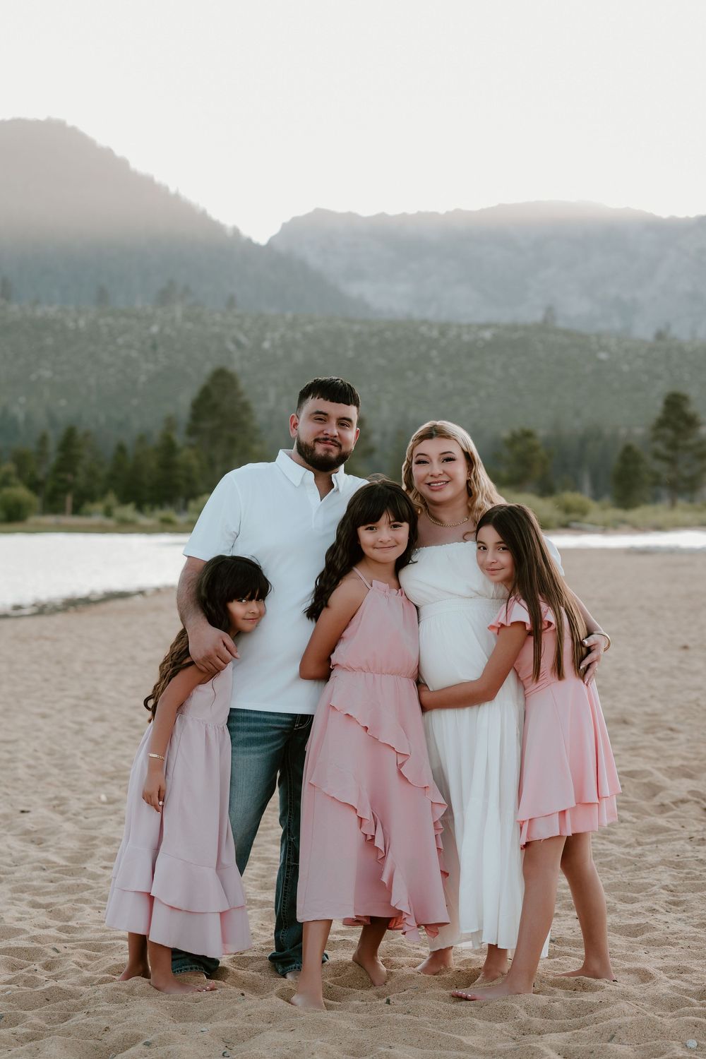 Family of five posing on sandy beach with mountain lake backdrop, parents with three girls in pink dresses.