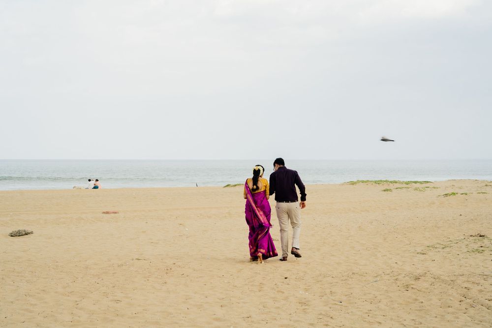 Couple walking hand in hand on a sandy beach, woman in vibrant pink saree, man in dark jacket and beige pants.