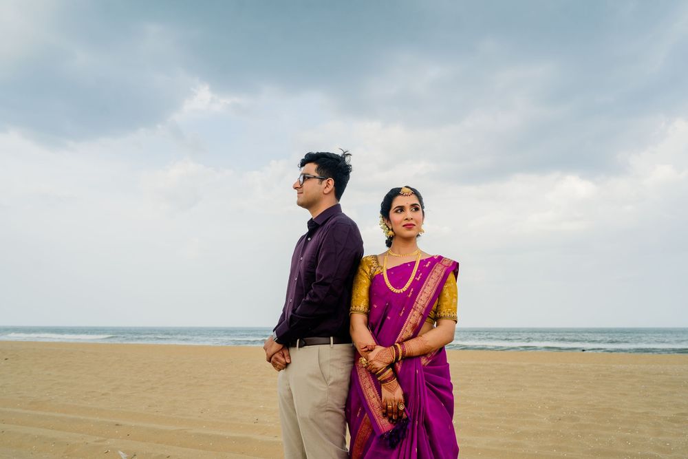 Indian couple in traditional wedding attire standing back-to-back on a beach, woman in purple saree, man in maroon shirt.