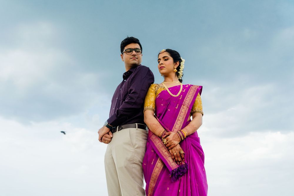 Indian couple in traditional attire, woman in pink saree and man in purple shirt, posing against cloudy sky.