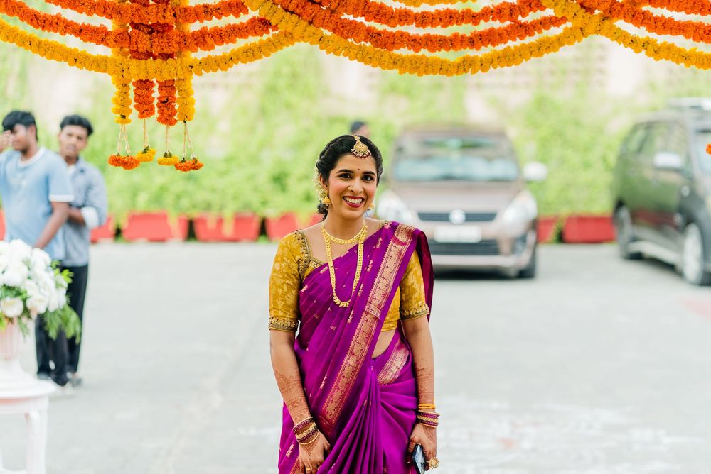 Woman in vibrant purple and gold silk saree smiles under marigold floral arch at Indian wedding.