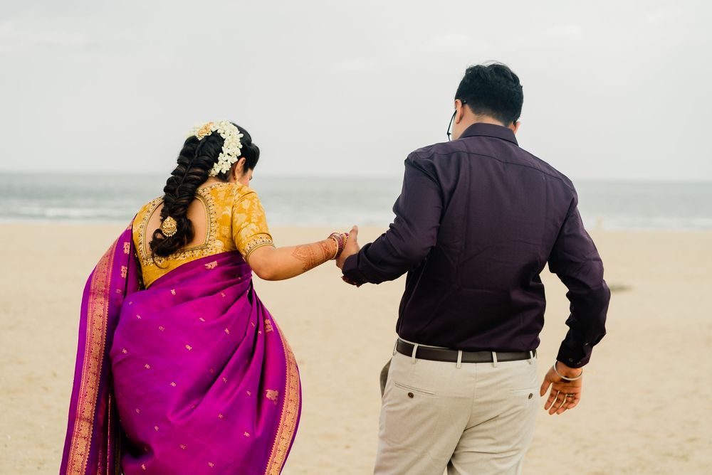 South Indian bride in purple silk saree and groom walking hand in hand on a sandy beach.