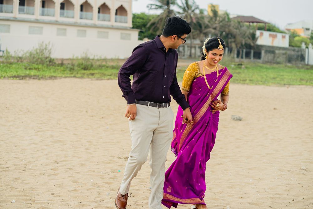Indian couple in traditional attire walking on sandy beach, woman in purple saree with gold jewelry.