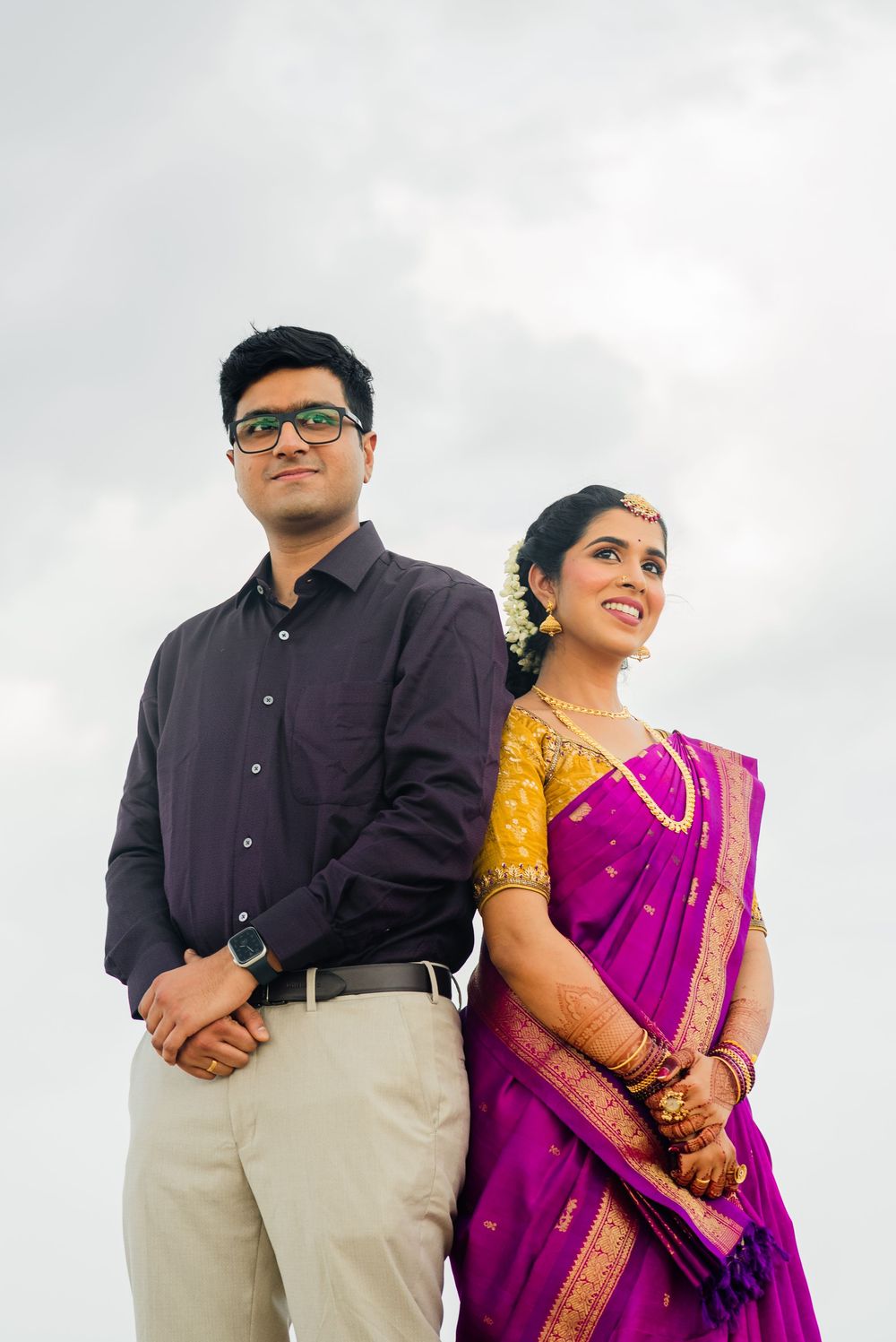 Indian couple in traditional attire, bride in purple saree with gold blouse, groom in purple shirt, posing against cloudy sky.