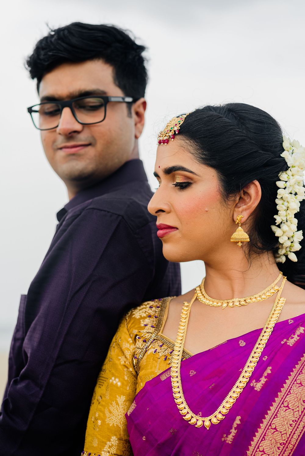 Indian bride in purple saree with gold jewelry and groom in dark shirt posing back-to-back at their wedding ceremony.