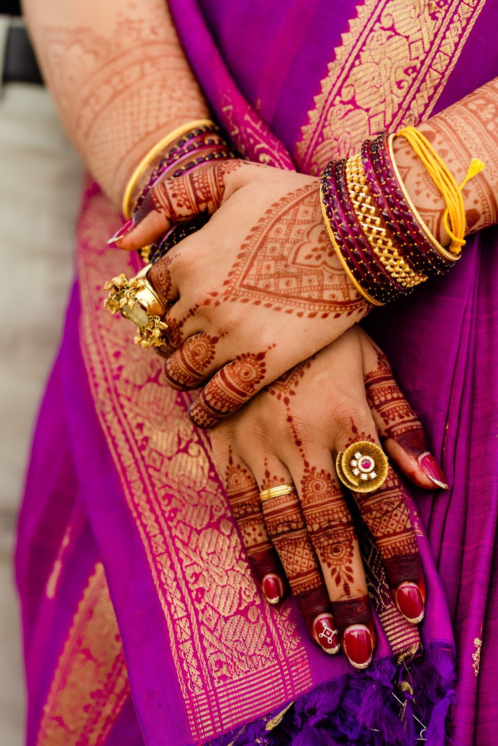 Indian bride's hands adorned with intricate henna mehndi designs, gold rings, and colorful bangles against a purple silk saree.