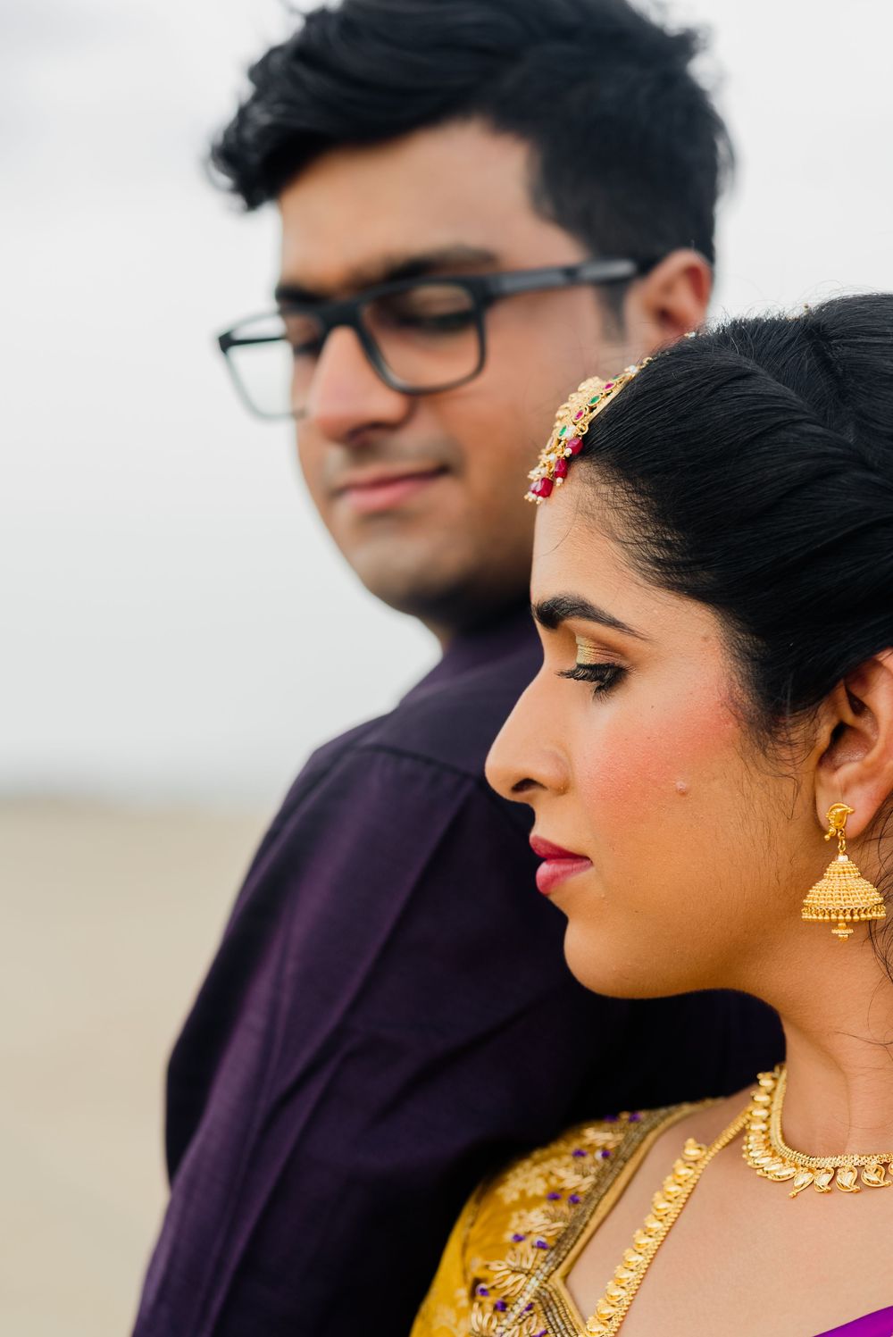 Indian bride in gold jewelry and yellow saree with groom in purple shirt during traditional wedding portrait.