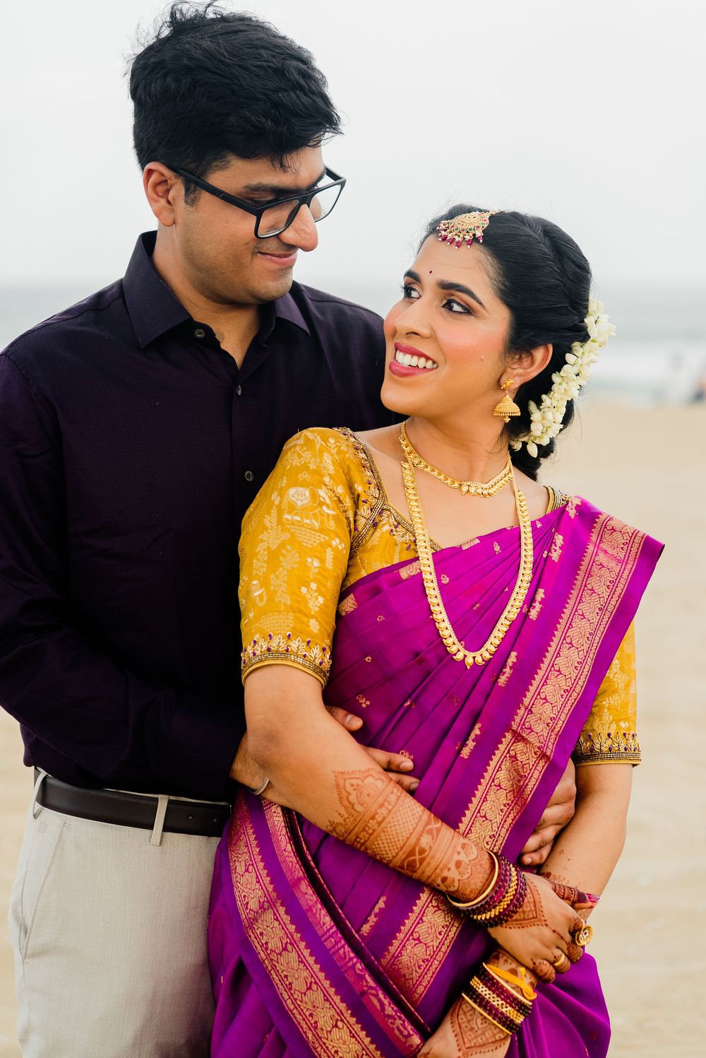 Indian couple in traditional attire on beach, bride in purple saree with yellow blouse and gold jewelry, groom in dark shirt.