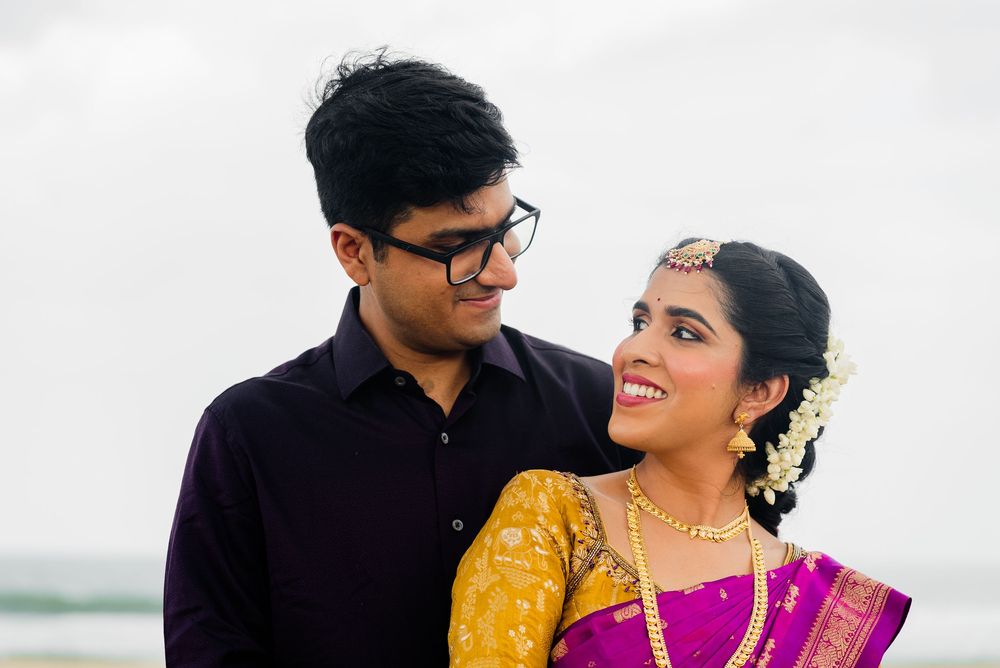 Indian couple in traditional wedding attire smiling at each other on a beach, bride in purple saree with gold jewelry.