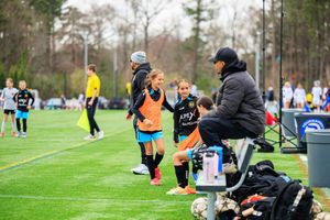 Youth soccer players in orange vests receive coaching on a green field during a competitive match.