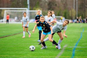 Young girls compete in a youth soccer match on a green turf field, with players dribbling and defending the ball.