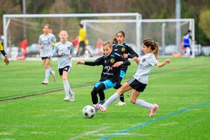 Young girls compete in a youth soccer match on a green turf field, with players dribbling and defending the ball.