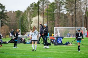 Female soccer players practice on a green field with a coach pointing directions near a goal post.