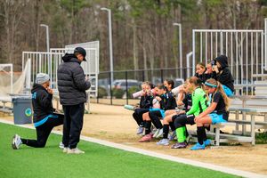 Soccer coach kneeling to address young female players sitting on bleachers during a youth soccer game.