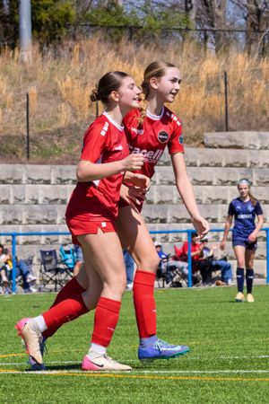 Two female soccer players in red uniforms celebrating together on turf field during a youth soccer game.