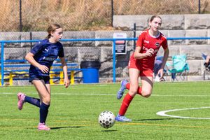 Two female soccer players competing for the ball on a turf field, one in red and one in navy uniform.