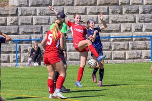 Female soccer players compete for the ball during an outdoor match on a sunny day at a stone-walled field.