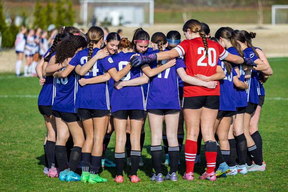 Girls soccer team huddles together in blue uniforms on green field during a match.