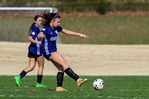 Two girls playing soccer on a grass field, one dribbling the ball in blue uniforms during a youth match.