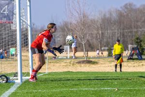 Female goalkeeper in red uniform deflects soccer ball near goal post on sunny day outdoor match.