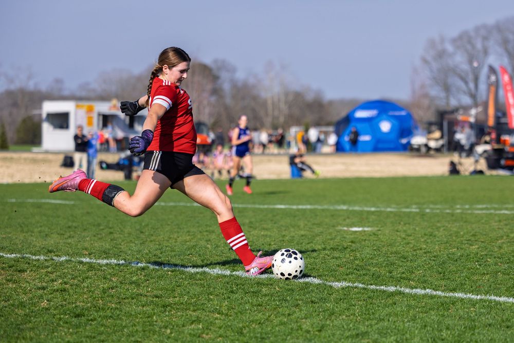 Young female soccer player in red uniform kicking ball on green field during outdoor youth match.