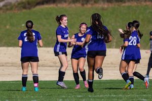 Girls youth soccer team in purple uniforms celebrating together on a sunny outdoor field during a match.