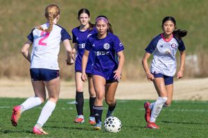 Young girls playing competitive youth soccer match on green field, players in purple and white uniforms chasing ball.