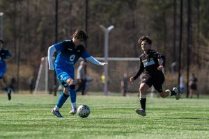 Two youth soccer players compete on a green turf field, one in blue dribbling the ball while another in black defends.