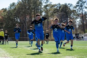 Youth soccer players in blue uniforms celebrate a goal on a green field with trees in the background.