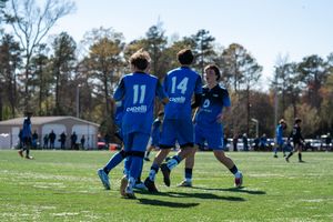 Youth soccer players in blue Capelli jerseys celebrate on a sunny green field during a competitive match.