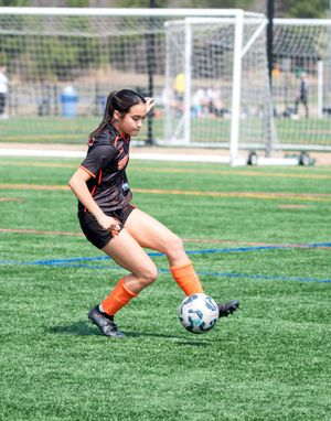 Young female soccer player in black and orange uniform dribbling a ball on a green turf field.