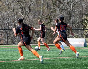 Female soccer players in orange uniforms compete on a green turf field during a sunny spring match.