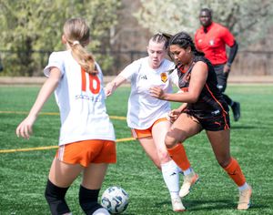 Female soccer players compete for the ball during an outdoor match on a green turf field in orange and white uniforms.