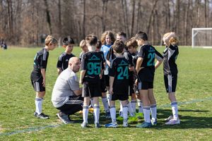 Youth soccer team in black jerseys huddles with coach on green field during sunny day practice or game.