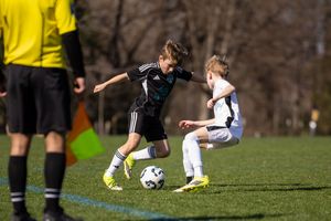 Two young soccer players compete for the ball during a youth match on a green field, with a referee visible nearby.