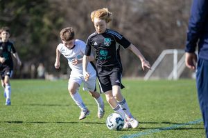 Young boys playing competitive youth soccer match on green field, player in black dribbling ball past opponent.