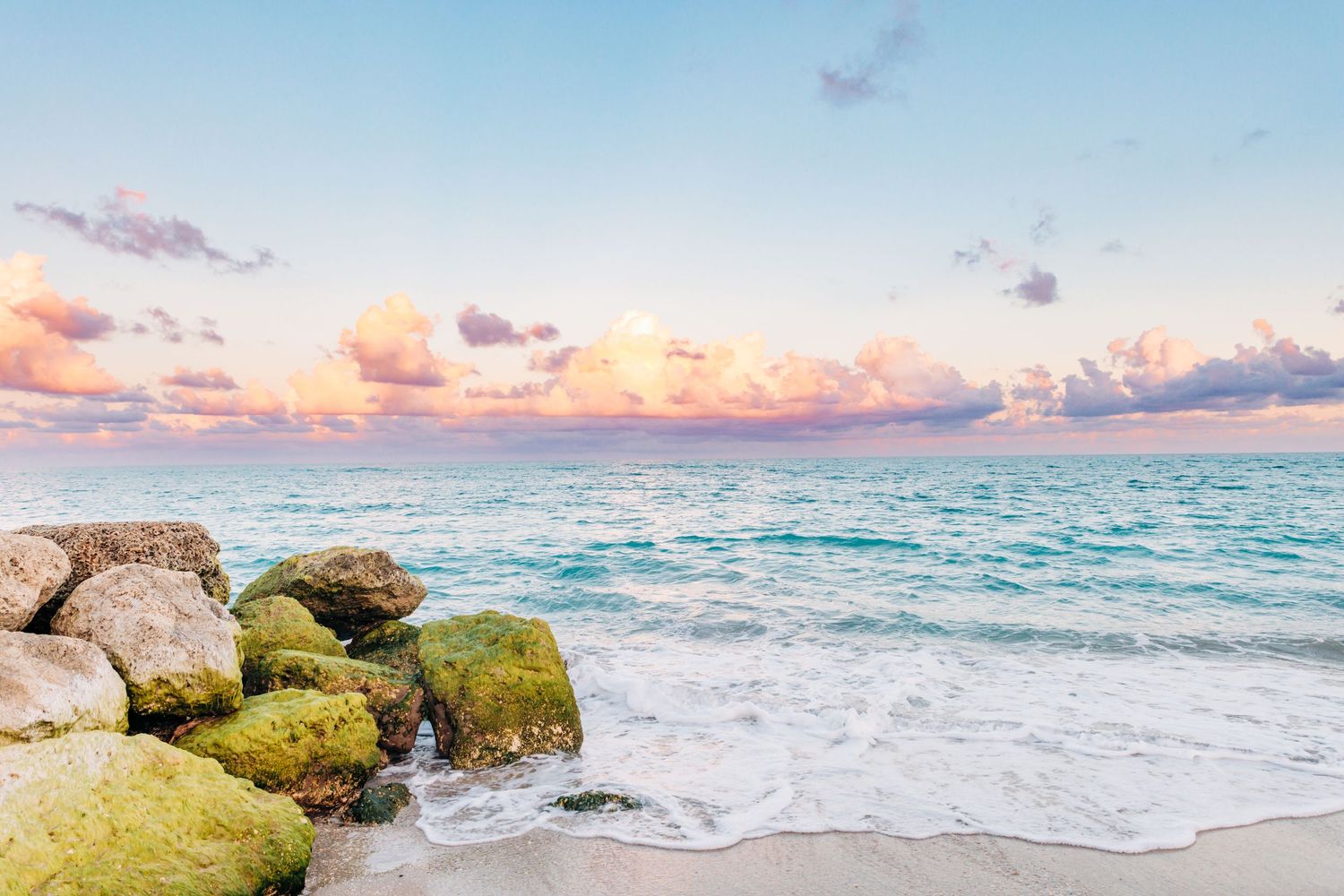 Tropical beach at sunset with moss-covered rocks, turquoise waves, white sand, and pastel pink clouds over the ocean in palm beach, beach.