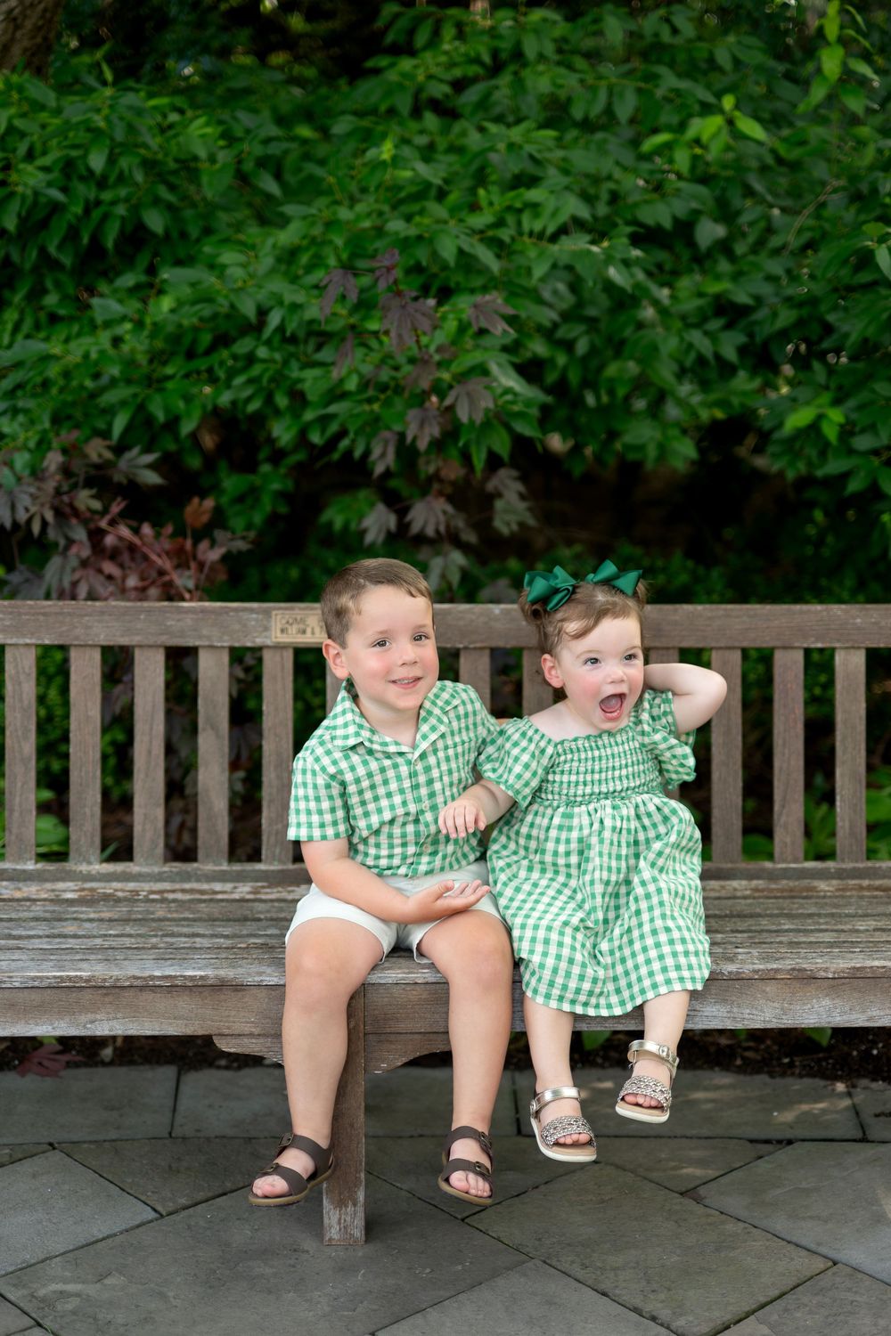Two young children in matching green gingham outfits sitting on a wooden bench outdoors.