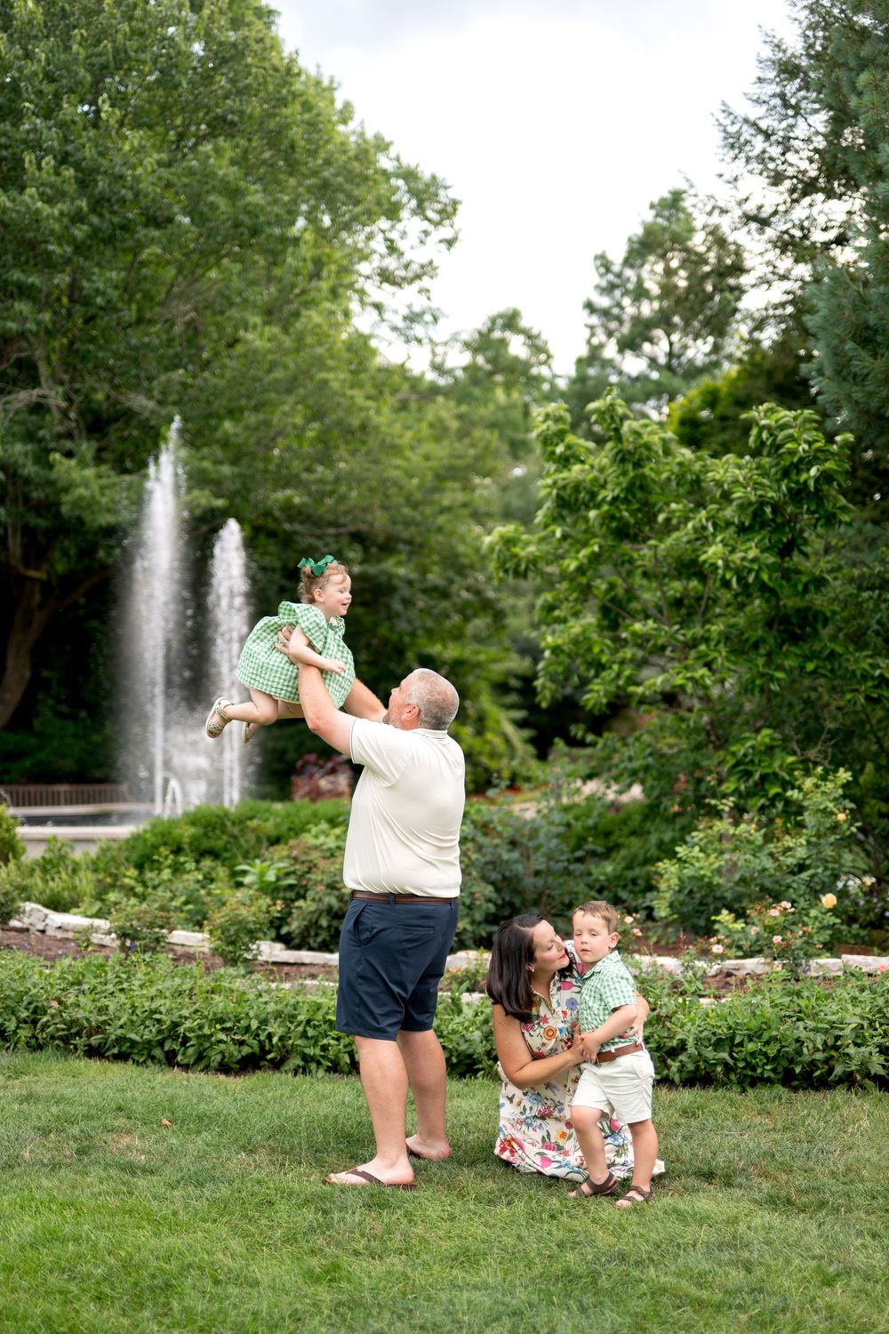 Family of four playing in a lush garden park with a fountain, dad lifting daughter while mom holds son.
