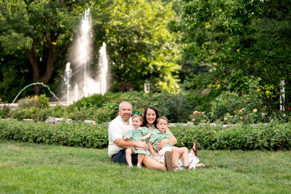 Family of four sitting on green grass in front of a fountain and lush garden, wearing coordinating green and white outfits.