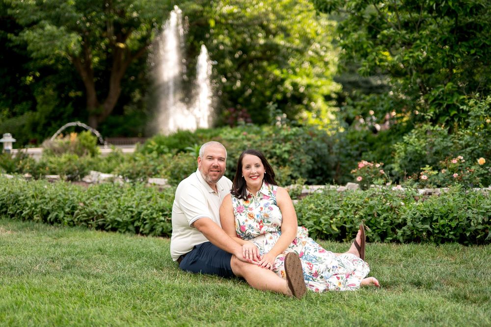 Couple sitting on green grass in a garden with a fountain and lush trees in the background.