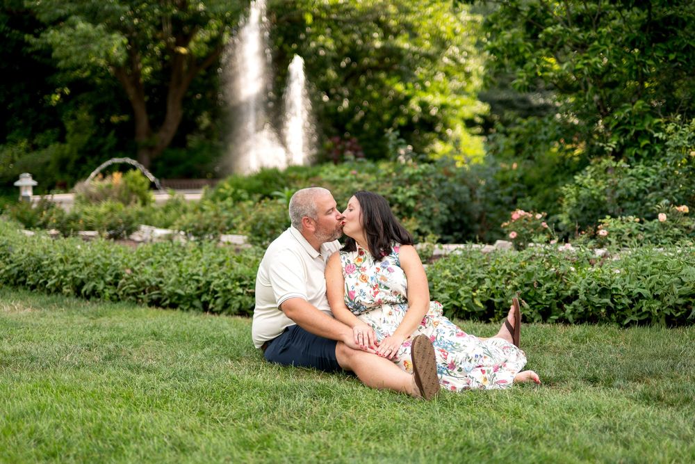 Couple sharing a romantic kiss while sitting on grass in a garden with a fountain backdrop.